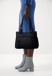 Sac tote noir en nylon, avec des poignées doubles et une poche zippée. Le sac est porté par une personne vêtue d'une robe bleu côtelée et de bottes grises.