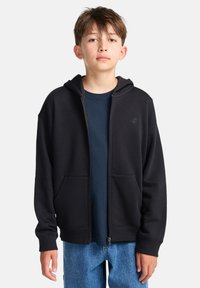 Felpa nera con zip, tasche laterali, polsini a costine e cappuccio. Indossata sopra una maglietta blu scuro; il modello indossa jeans azzurri.