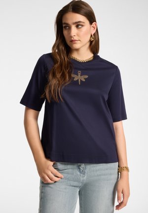 Femme aux longs cheveux bruns portant une chemise bleu marine avec un motif libellule doré et un jean bleu clair, accessoirisée de bijoux en or.