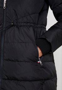 Chaqueta negra tipo plumas con acabado texturizado, que cuenta con cintura elástica, bolsillos con cremallera y puños de canalé. Herrajes de cremallera plateados visibles.