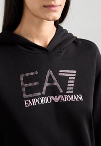 Fekete kapucnis felső, ezüst és rózsaszín strasszokkal díszített "EAT" felirattal, alatta pedig fehér színnel nyomtatva az "EMPORIO ARMANI" felirat.