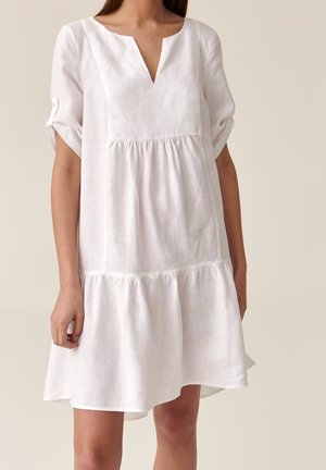 Kobieta ubrana w luźną, białą sukienkę z krótkim rękawem, dekoltem w kształcie litery V i marszczonymi warstwami, stojąca na tle gładkiego beżowego tła.