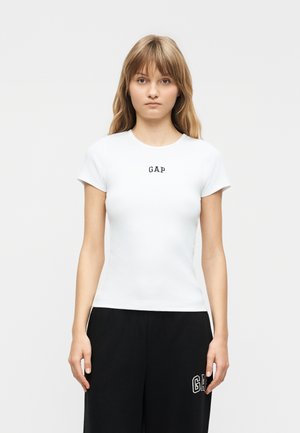 Joven con cabello lacio castaño claro lleva una camiseta blanca ajustada de GAP y pantalones negros sueltos de GAP, de pie contra un fondo liso.