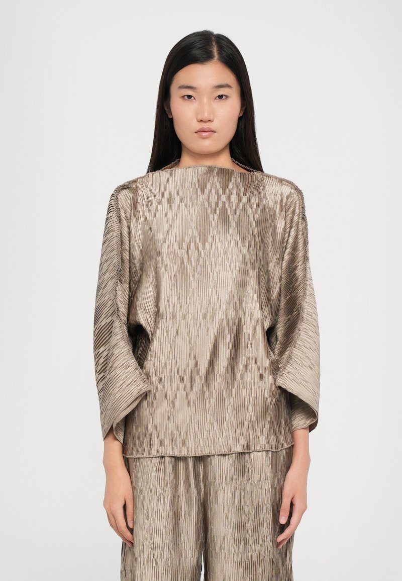 Light beige, pleated top with a relaxed fit, wide sleeves, and a smooth texture. Features vertical lines and a minimalist design.