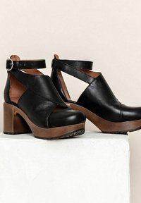 Zwarte leren klompen met een uitsnijdontwerp, enkelbandje en bruine houten zool. Stevige blokhak en geprofileerde zool voor grip.