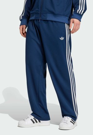 Personne portant un pantalon de survêtement bleu Adidas avec des bandes blanches et un logo, associé à des baskets blanches Adidas avec des bandes noires.