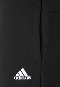 Schwarze Baumwoll-Sweatpants mit seitlicher Tasche und weißem Adidas-Logo unten links. Weiche Textur mit klassischem Design.