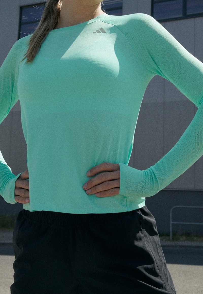 Mintgrön långärmad träningsskjorta med en åtsittande design, texturerade ärmar och logotyp på bröstet. Känns tillsammans med svarta shorts.
