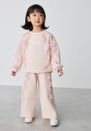 Junges Kind mit schwarzen Haaren, das einen hellrosa gerüschten Pullover und Hose trägt, steht auf einem grauen Boden vor einem schlichten hellen Hintergrund.