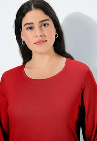 Červená košile s dlouhým rukávem a kulatým výstřihem, vyrobená z hladké tkaniny s jemnou strukturou, která má přiléhavý střih.