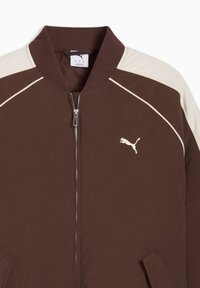 Καφέ ζακέτα Puma με φερμουάρ, με μπεζ πλαϊνά στους ώμους και λευκό λογότυπο Puma στο στήθος, με ριμπ γιακά και πλαϊνές τσέπες.