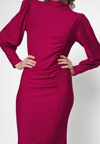 Femme portant une robe magenta ajustée à manches longues avec un détail froncé sur le côté et des épaules bouffantes, les mains posées sur les hanches.