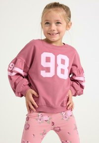 Sweatshirt rose vif avec le numéro blanc "98" sur le devant et les manches, manches longues bouffantes ; associé à des leggings roses avec motifs floraux.