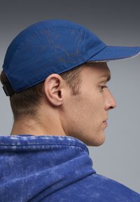 Blå baseballcap med flad skygge, med et diskret design på siden, båret af en person i en struktureret blå hættetrøje.