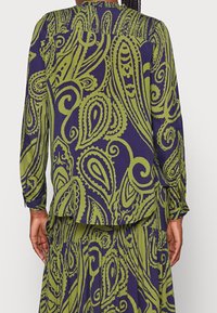 Pitkähihainen pusero ja hame, joissa on suuria vihreitä paisley-kuvioita tumman violetilla taustalla, henkilö pää selin esillä.