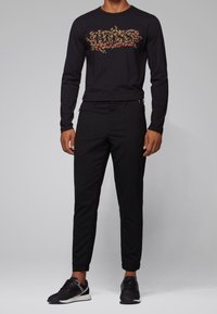 Czarna koszula z długim rękawem z wzorzystym logo „BOSS” w czerwieni i złocie. Do tego dopasowane czarne spodnie i czarne sportowe buty.