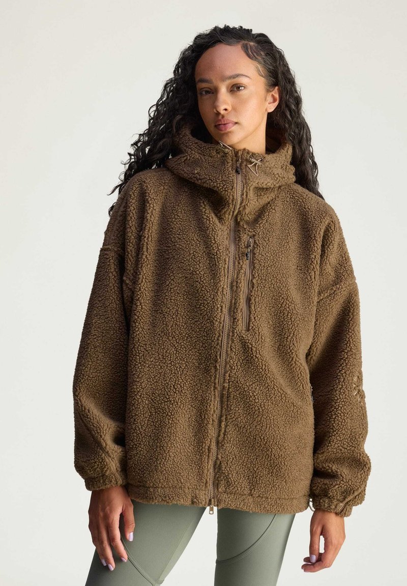 Brown fleece jacket with a high collar, full front zipper, and side pockets. Features a textured, soft exterior and oversized fit.