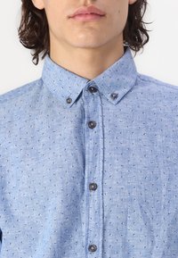Chemise à manches longues bleu clair en tissu avec de petits motifs à pois foncés. Elle présente un col classique et des boutons marron sur le devant.