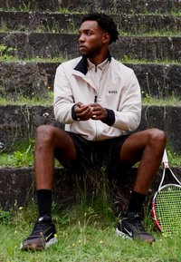 Hellbeige Sportjacke mit schwarzen Akzenten, schwarze Shorts und Turnschuhe. Tennisschläger daneben, sitzt auf Steinstufen mit Gras.