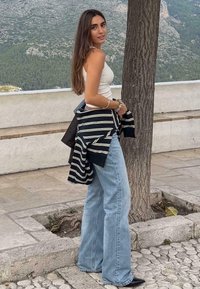 Wit tanktop gecombineerd met lichtblauwe uitlopende jeans. Gestyled met een zwart-wit gestreepte jas losjes over de schouder gedragen. Buitenshuis setting.