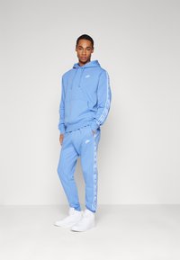 Zila Nike kapuce ar kangaroo kabatu, balts logo un saskanīgi bikses. Abiem ir balti sānu svītri. Valkā ar baltiem augstajiem čībām.