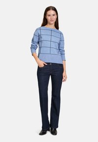 Blauer, langärmeliger Pullover mit schwarzem Karomuster, kombiniert mit dunklen Jeans und schwarzem Schuhwerk. Der Pullover hat einen schmalen Rundhalsausschnitt.