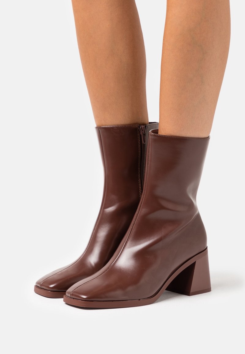 Bottes en cuir marron mi-mollet avec talons blocs et fermetures éclair latérales portées sur des jambes nues sur fond blanc.