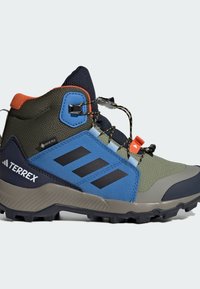 Τα μποτάκια πεζοπορίας Terrex διαθέτουν έναν συνδυασμό υλικών σε ελιά, μπλε και γκρι, υφασμάτινη υφή, ψηλό σχέδιο στον αστράγαλο και μαύρη σόλα με πρόσφυση.