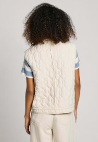 Prošívaná smetanová vesta s kulatým výstřihem a bez rukávů, s hladkou texturou a jemnými švy podél švů.