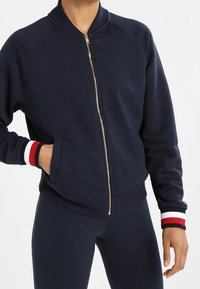 Námořnicky modrá bomber bunda na zip s žebrovaným límcem, bílými a červenými proužkovanými manžetami a bočními kapsami. Měkký materiál, pohodlný střih.