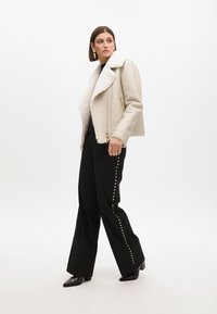 Giacca in finta pelliccia con colletto, dettagli in oro sulla cerniera, abbinata a pantaloni neri a gamba larga decorati con borchie argentate lungo il lato. Stivali neri a punta.