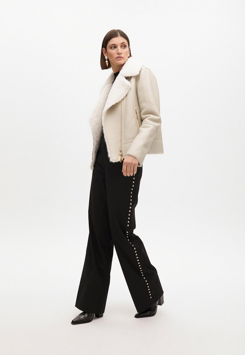 Giacca in finta pelliccia con colletto, dettagli in oro sulla cerniera, abbinata a pantaloni neri a gamba larga decorati con borchie argentate lungo il lato. Stivali neri a punta.