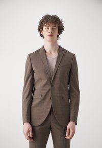 Costum carouri maro cu rever zimțat, închidere cu un singur nasture și două buzunare frontale; purtat cu un tricou tricotat bej deschis dedesubt.