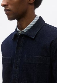 Σκούρο τζιν τζάκετ με κλείσιμο μπροστά με κουμπιά, δύο τσέπες στο στήθος και πουκάμισο με γιακά με ανοικτές μπλε και λευκές ρίγες από κάτω.