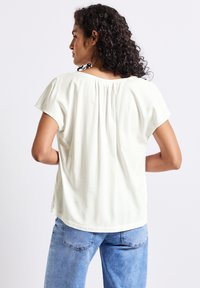 Blusa de manga corta en color crema, con un corte relajado, detalle de pliegue en la parte posterior y textura suave. Combinada con denim azul claro.