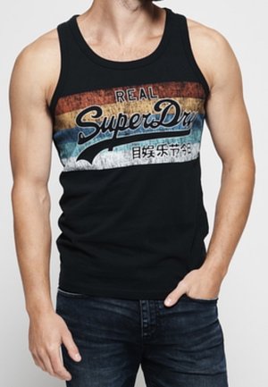 Hombre con camiseta sin mangas negra con rayas retro rojas, amarillas y azules y texto "Real SuperDry", combinado con vaqueros oscuros, mano en el bolsillo.