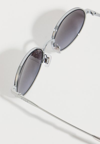 Ασημένια αεροπόρα γυαλιά ηλίου με μεταλλικό σκελετό και σκούρους φακούς, με ρυθμιζόμενα μαξιλαράκια μύτης, τοποθετημένα σε λευκή επιφάνεια σε γωνία.