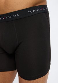 Μαύρα ανδρικά μποξεράκια Tommy Hilfiger φορεμένα σε ανδρικό κορμό, με λογότυπο στη μέση και λεία υφή υφάσματος.