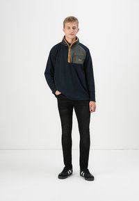 Pullover in pile blu navy con colletto con zip, taschino verde sul petto e dettagli in zip arancioni, abbinato a jeans neri slim-fit e sneakers nere.