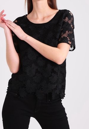 Mujer con una blusa negra de encaje floral de manga corta combinada con pantalones negros, de pie con las manos juntas cerca de la cara y el pecho.