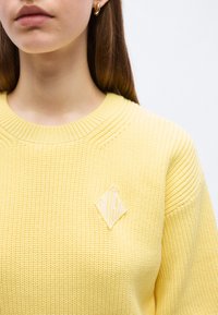Γυναίκα που φοράει κίτρινο πλεκτό πουλόβερ με λευκή λογότυπη ετικέτα σε σχήμα ρόμβου στο στήθος και μικρό χρυσό κρίκο στο αυτί.