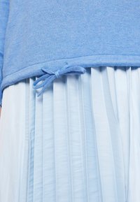 Ljusblå stickad tröja med en justerbar knytning vid nederkanten, matchad med en ljusblå plisserad kjol med vertikala veck.