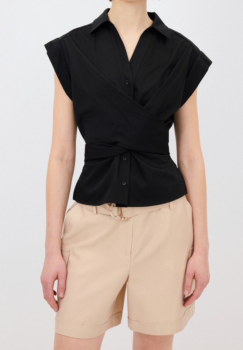 Femme portant un chemisier noir à manches courtes avec boutons et un short beige longueur genou avec poches et passants pour ceinture.