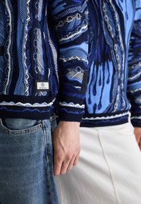 Pull en tricot bleu et marine avec des motifs de vagues complexes, doté de poignets côtelés et d'un patch logo, associé à un jean en denim.