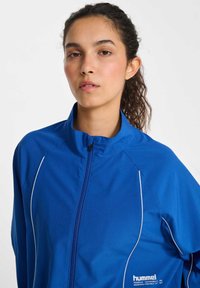 Junge Frau mit lockigem Haar im Pferdeschwanz trägt eine blaue Hummel-Sportjacke mit Reißverschluss und weißen Paspeln vor einfarbigem Hintergrund.