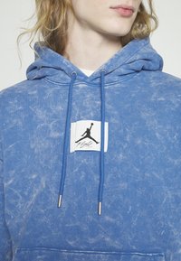 Sudadera azul hecha de un tejido suave y texturizado, con un gran parche de logo blanco que presenta una silueta negra y el texto "Flight", y cordones con puntas plateadas.