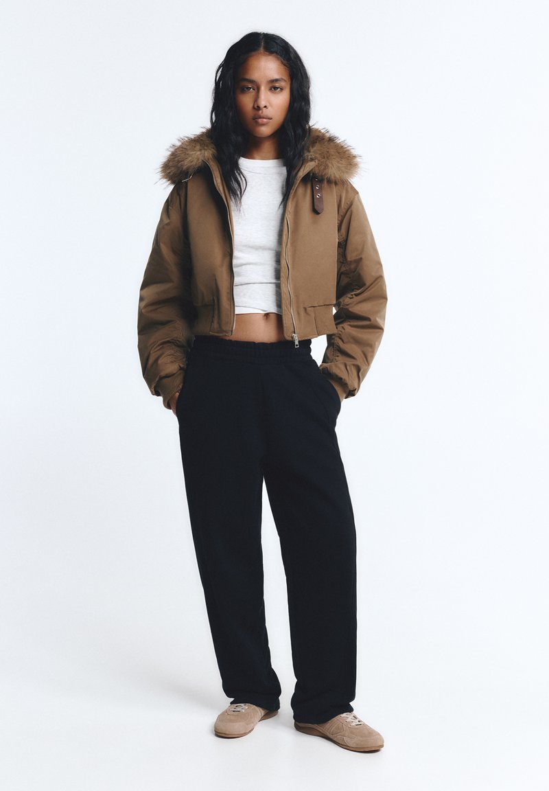 Bruine cropped jack met een met bont gevoerde capuchon, grijze crop top, zwarte broek en beige schoenen. Het jack heeft een ritssluiting en elastische manchetten.