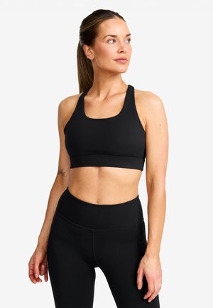 Fit kvinna i svart sportbh och leggings ser åt sidan, stående mot en enfärgad vit bakgrund.