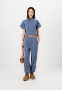 Gekürztes blaugraues Sweatshirt mit kurzen Ärmeln, kombiniert mit passenden lockeren Sweatpants. Das Model hält eine braune Umhängetasche. Braune Plateauschuhe.