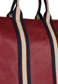 Rode leren tas met textuur, voorzien van marineblauwe, witte en oranje gestreepte stoffen hengsels tegen een witte achtergrond.